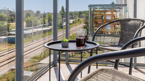 A balcony with a view of railway tracks, a small table with a plant and two glasses of red wine, and wicker chairs.
