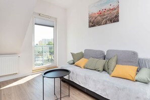 Un salon moderne avec un canapé gris, de nombreux coussins, une petite table noire et une grande fenêtre donnant sur des immeubles et de la verdure.