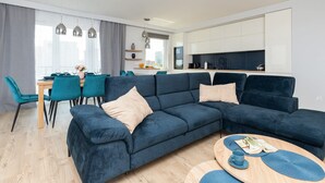 Ein modernes Wohnzimmer mit einem dunkelblauen Ecksofa, Türkis-Farbenen Sesseln und einem runden Couchtisch. Im Hintergrund ist ein Essbereich mit einem Holztisch und Stühlen zu sehen.