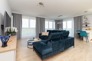 Ein modernes Wohnzimmer mit einem dunkelblauen Ecksofa, einem Holztisch und einem Essbereich mit türkisfarbenen Stühlen und einem weißen Tisch.