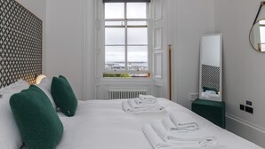 Hotellihuone, jossa on suuri sänky, vihreät tyynyt, ikkunasta avautuva näkymä kaupunkiin, patteri ja peili seinällä.