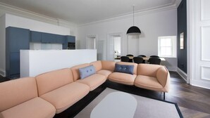 A modern living room with a beige sectional sofa, a dining area with a black table and chairs, and a kitchen with blue cabinets. - Ranol (Isle of Lewis)
