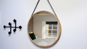 A round mirror with a wooden frame hanging from a cord. It reflects a room with a bed, a window, and a wall-mounted light fixture. - Seaforth North (Isle of Lewis)
