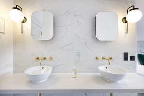 Un baño moderno con dos lavamanos blancos tipo sobremesa, grifos dorados y espejos empotrados con luces redondas.