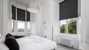 Une chambre avec un grand lit, du linge de lit blanc, un store foncé à la fenêtre, un radiateur et une vue sur la verdure à l’extérieur.