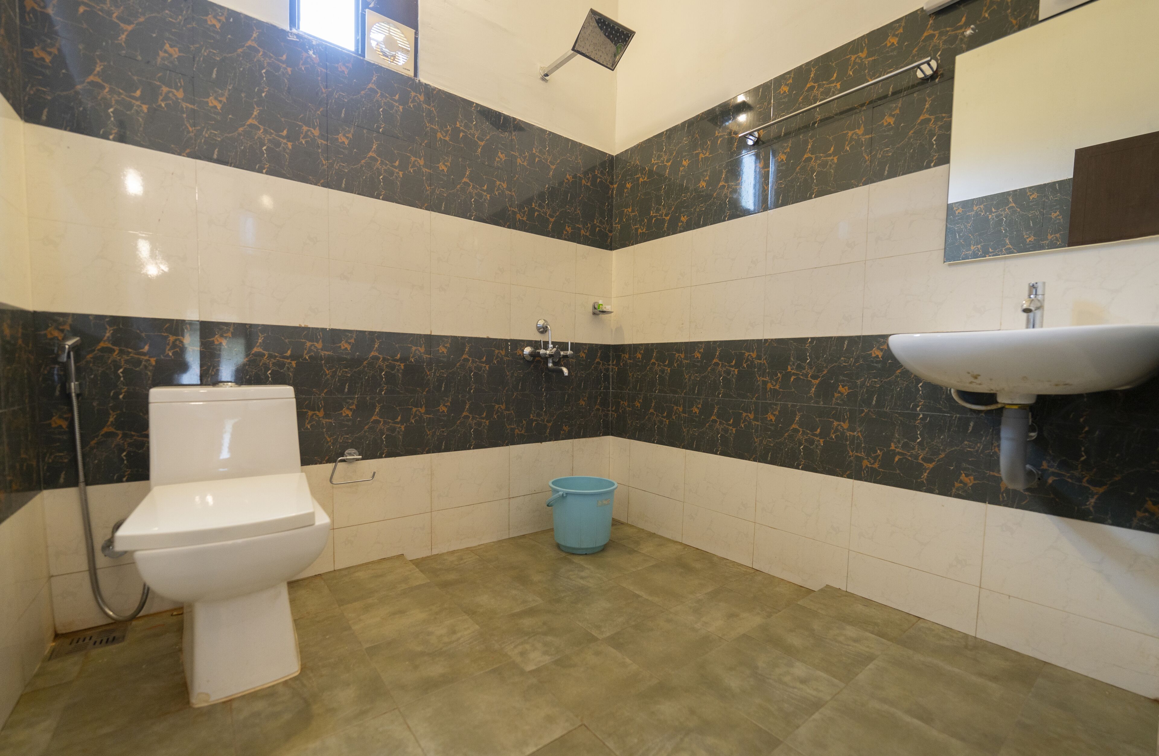 A bathroom with a toilet, a blue bucket, a shower, and a sink.