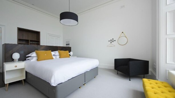 Un dormitorio moderno con una cama gris, ropa de cama blanca, una lámpara colgante negra, una silla negra y un reposapiés amarillo.