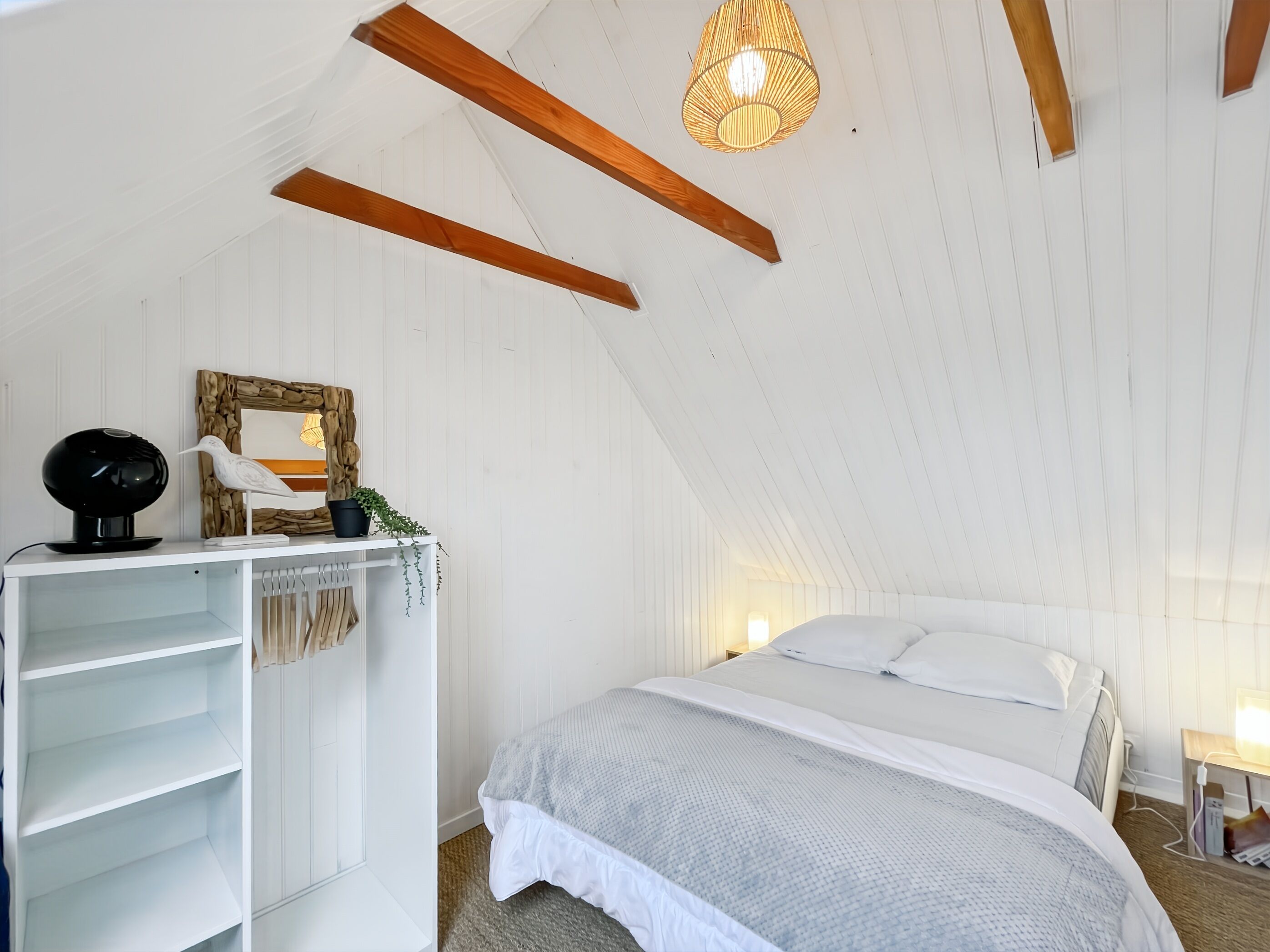 A bedroom with a sloped ceiling, a bed with white bedding, a white wardrobe, a small table with a lamp, and a wall-mounted shelf with decorative items.