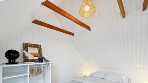 Et soveværelse med skrå væg, seng med hvide sengetøj, et hvidt klædeskab, et lille bord med lampe, og en vægmonteret hylde med pynt.