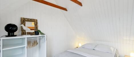 A bedroom with a sloped ceiling, a bed with white bedding, a white wardrobe, a small table with a lamp, and a wall-mounted shelf with decorative items.