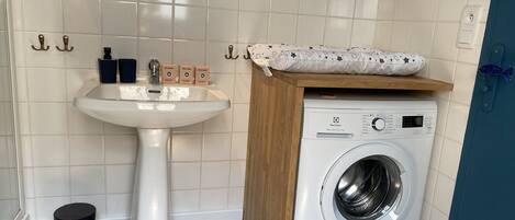 A compact bathroom with a pedestal sink, a washing machine, and a small trash bin.
