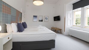 A modern bedroom with a large bed, a bedside table, a small table, a TV, and a window with a view of greenery. - Ranol East (Isle of Lewis)