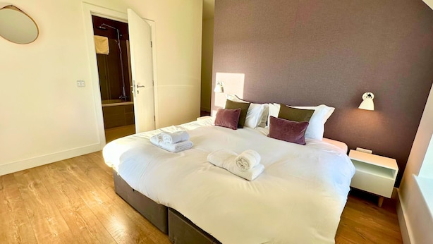A neatly made bed with white linens and rolled towels, a bedside table with a lamp, and a wooden floor. - Northstar - 4 Bed Luxury Apartment (Wick)