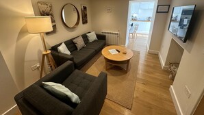 A modern living room with a sectional sofa, a round coffee table, and a floor lamp. There is a kitchen visible through an open doorway. - Fowey - Crimpers Cottage (Fowey)