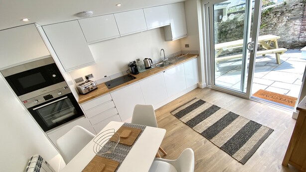 A modern kitchen with white cabinets, a built-in oven, and a coffee maker. There is a dining area with a table and chairs, and a view of the outside through a glass door. - Fowey - Crimpers Cottage (Fowey)