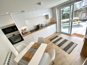 A modern kitchen with white cabinets, a built-in oven, and a coffee maker. There is a dining area with a table and chairs, and a view of the outside through a glass door. - Fowey - Crimpers Cottage (Fowey)