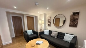 A modern living room with a round wooden coffee table, two black sofas with light-colored cushions, a round mirror, and a painting on the wall. - Fowey - Crimpers Cottage (Fowey)