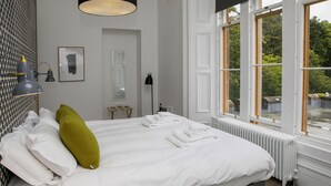 En snyggt bäddad säng med vita lakan och en grön kudde, ett element och ett fönster med utsikt över grönområden.