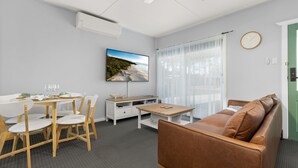 A modern, well-lit room with a dining area, a sofa, a TV, and a large window with sheer curtains. - Wanderer by Experience Jervis Bay (Sanctuary Point)