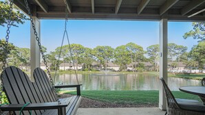 A covered patio with a swing seat and wicker table, overlooking a lake and residential area. - Seascape Villa 92F *Beach access and multiple pools! (Miramar Beach)
