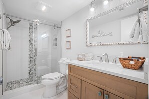 Une salle de bain moderne dotée d’une douche à parois de verre, de toilettes blanches, d’un meuble-lavabo en bois avec des tiroirs et d’un grand miroir agrémenté d’un cadre décoratif.