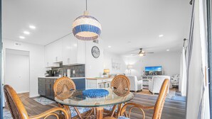 A modern kitchen with white cabinets, a dining area with a glass table and wicker chairs, and a living room with a sofa and a television. - Seascape Villa 92F *Beach access and multiple pools! (Miramar Beach)