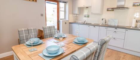 Una cocina moderna con armarios blancos, una mesa de comedor de madera dispuesta con platos azules y copas de vino, y un reloj en la pared.