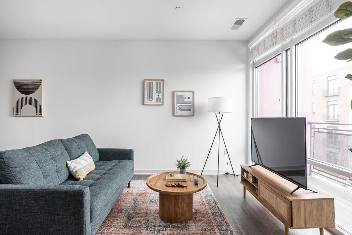 Ein modernes Wohnzimmer mit einem grauen Sofa, einem Holztisch, einem Fernseher auf einem Holzschrank und gerahmten Kunstwerken an der Wand.