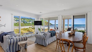 A spacious room with a large dining table and chairs, a sofa set, and a TV. The room has large windows offering a view of the beach and ocean. - Myponga Dunes - 1 Hewett Crescent, Myponga Beach (Myponga Beach)