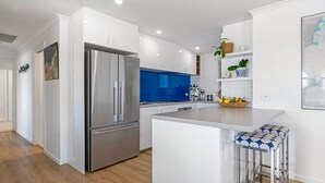 A modern kitchen with white cabinetry, a stainless steel refrigerator, a blue backsplash, and a small island with a bar stool. - Myponga Dunes - 1 Hewett Crescent, Myponga Beach (Myponga Beach)