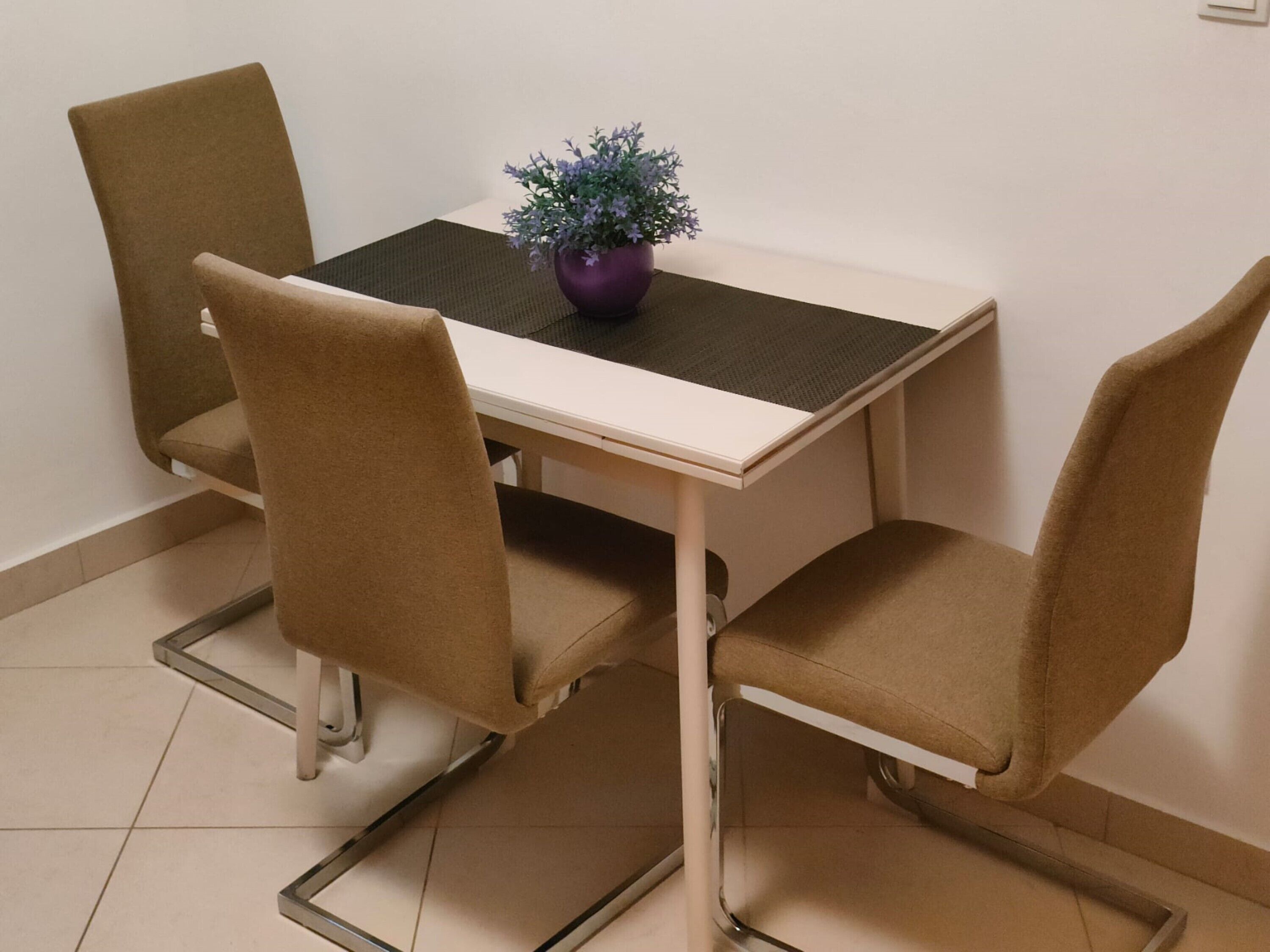 A small dining area with a white table and four brown chairs. There is a purple plant on the table.