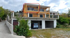 A two-story building with a balcony, a car parked in the driveway, and a small garden area.