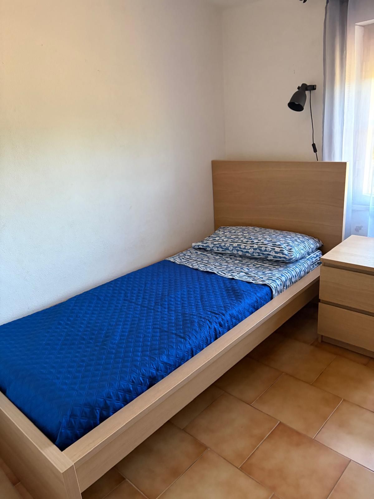 A single bed with a wooden frame and a blue quilted mattress cover, a bedside table with drawers, and a wall-mounted lamp.