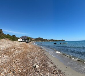 A pebble beach with a building and boats on the water.