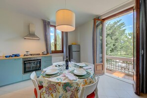A dining area with a table set for four, a kitchen with a stove and oven, and a view of trees through a balcony. - Casa Lia 2BR for 4 in Desenzano Parking & AC (Desenzano del Garda)