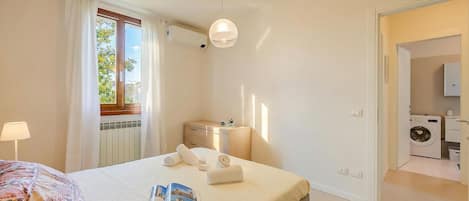 Una camera d'albergo con un letto, una finestra con tende, una lampada da comodino, un lampadario e un tavolino con una rivista, con vista su un'area lavanderia.