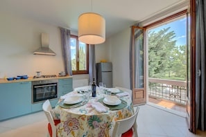 Uma sala de jantar com mesa posta para quatro pessoas, uma cozinha com fogão e forno, e uma varanda com vista para árvores.