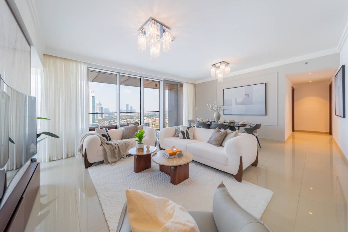 A spacious living room with a large window, a white sofa set, a coffee table, and a TV.
