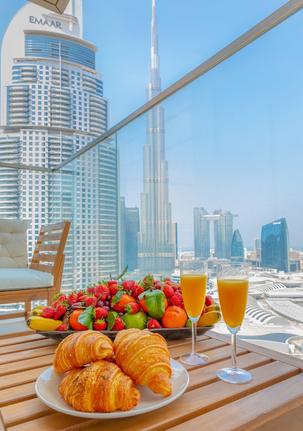 A breakfast table with croissants, fruit, and orange juice, overlooking a cityscape with tall buildings.