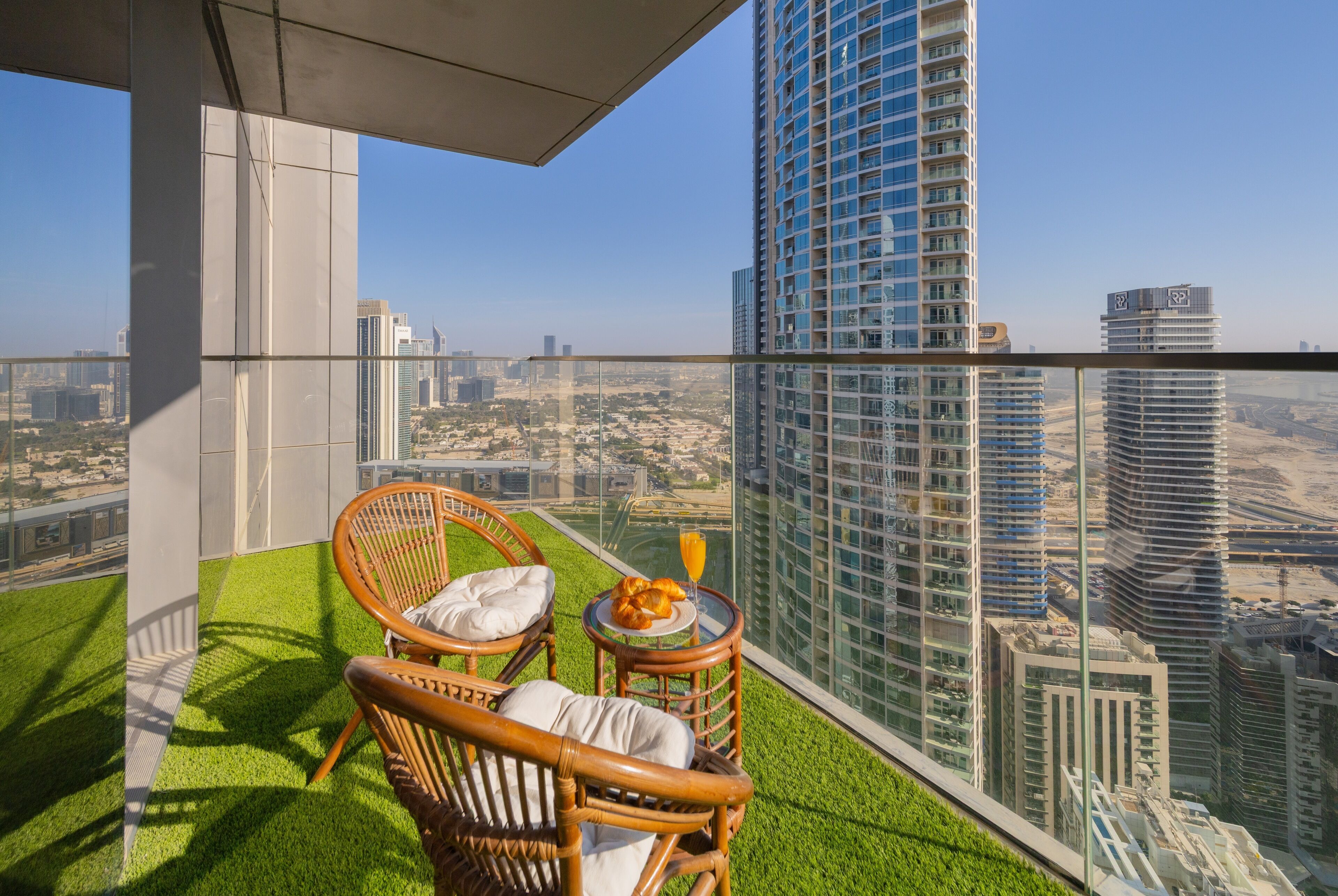 A balcony with artificial grass, wicker chairs, a small table with a glass of orange juice, and a city view.