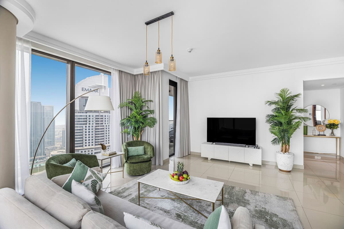 A modern living room with a sofa, coffee table, and TV, large windows with a city view, and potted plants.