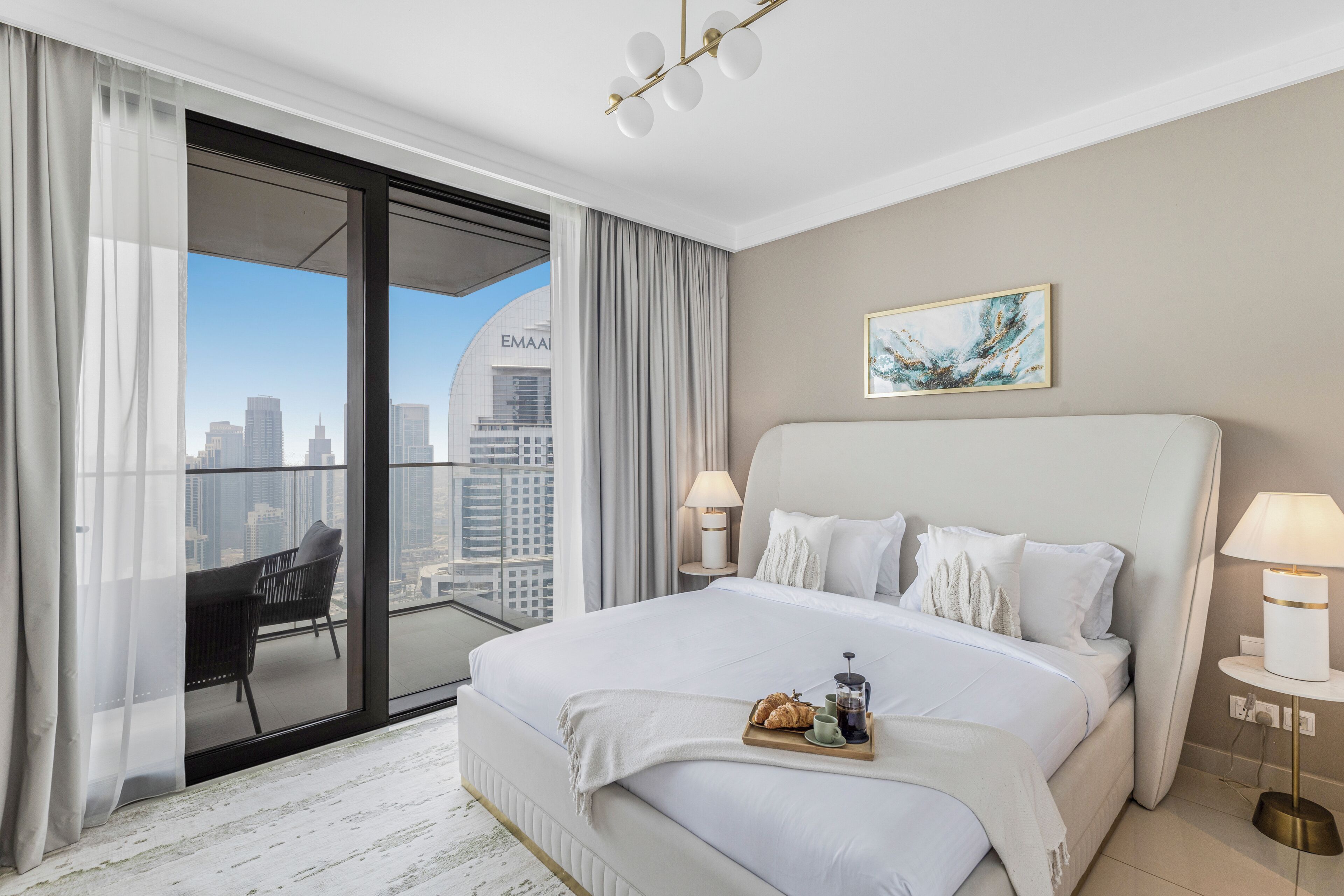 A modern hotel room with a large bed, a balcony with a city view, and a dining area with breakfast items.