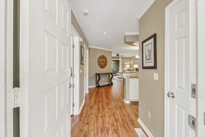 A hallway with white doors, wooden flooring, a clock on the wall, and a kitchen area in the background.