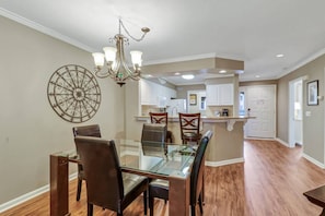 A dining area with a glass table and chairs, a chandelier, and a kitchen in the background.
