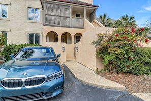 A blue BMW parked in front of a building with a balcony, a gated entrance, and a garden with red flowers.