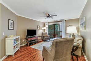 A living room with a flat-screen TV, a ceiling fan, a sofa, a chair, a bookshelf, and a balcony with a view of trees.