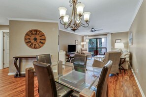 A dining area with a glass table and chairs, a large wooden clock on the wall, and a chandelier.
