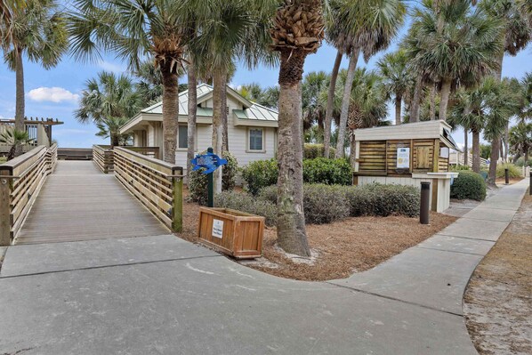 A wooden boardwalk leading to a building with a green roof, surrounded by palm trees. - 145 Colonnade Club Villa Beach Path, Pool, Hot Tub (Hilton Head Island)
