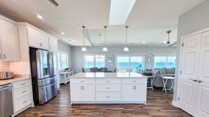 A spacious kitchen with a large island, white cabinetry, and a view of the ocean. - Mermaid Cove – Splash Into Relaxation (North Topsail Beach)