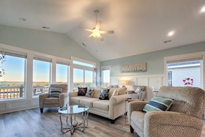 A spacious living room with a high ceiling fan, large windows, a sofa, armchairs, and a glass coffee table.